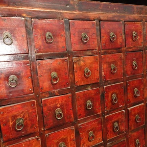 Op de afbeelding: Een vintage houten kast met meerdere kleine laden. De laden hebben messing ringgrepen en zijn in een dieprode kleur geverfd. De kast is gemaakt van donker hout en heeft een verweerde uitstraling.