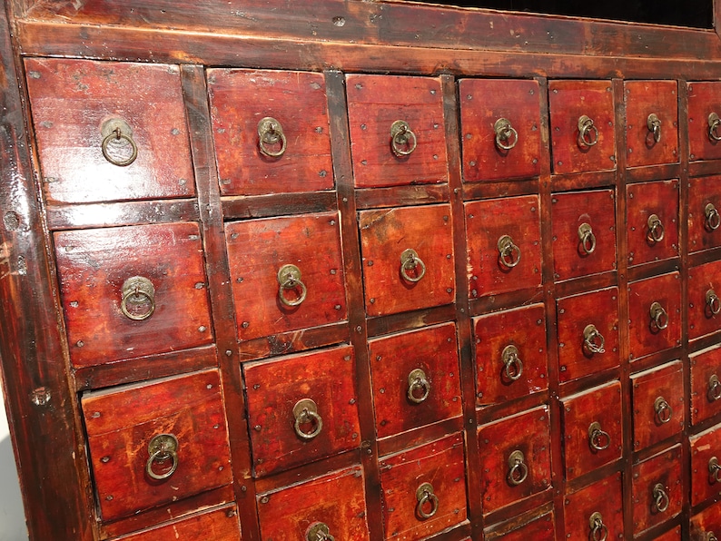 Op de afbeelding: Een vintage houten kast met meerdere kleine laden. De laden hebben messing ringgrepen en zijn in een dieprode kleur geverfd. De kast is gemaakt van donker hout en heeft een verweerde uitstraling.