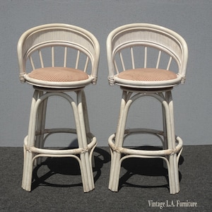 May include: Two white wicker bar stools with a checkered red and white cushion. The stools have a rounded back and a swivel seat.