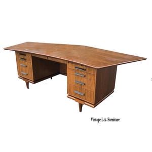 May include: A vintage wooden desk with a unique angular design. The desk features multiple drawers with silver handles and tapered legs. The desk is a warm brown color and has a large surface area.