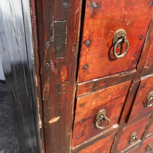 Op de afbeelding: Antiek houten kast met meerdere laden. De laden zijn rood geverfd en hebben een zwarte rand. Elke lade heeft een metalen ringvormige trekknop.