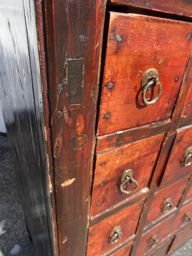 Op de afbeelding: Antiek houten kast met meerdere laden. De laden zijn rood geverfd en hebben een zwarte rand. Elke lade heeft een metalen ringvormige trekknop.