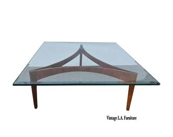 May include: A square glass-top coffee table with a wooden base. The base features a unique design with curved and angled wooden supports. The table has four tapered legs and is set against a white background. The text "Vintage L.A. Furniture" is at the bottom.