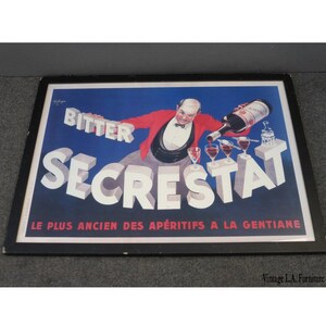 May include: A vintage framed poster advertising Secrestât Bitter, a French aperitif. The poster features a man in a red jacket pouring the drink into glasses. The text on the poster reads "Bitter Secrestât Le Plus Ancien Des Apéritifs A La Gentiane".