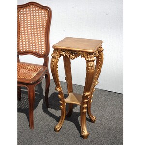 May include: A gold-colored, ornate plant stand with a square top and curved legs. The stand has decorative carvings and a small shelf. A wooden chair with a woven back is partially visible in the background.