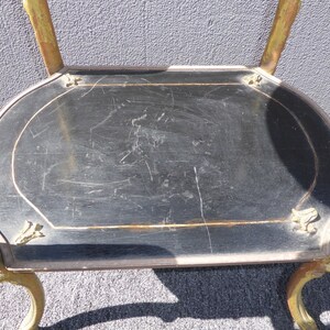 May include: A black and gold metal serving cart with a round top and ornate legs. The top is made of wood and has a black finish. The cart is in good condition, but shows signs of wear and tear.