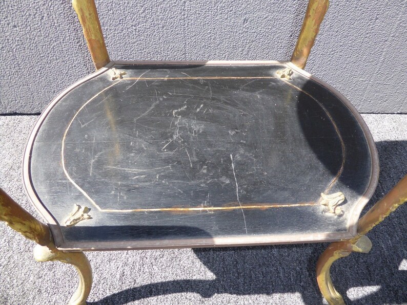May include: A black and gold metal serving cart with a round top and ornate legs. The top is made of wood and has a black finish. The cart is in good condition, but shows signs of wear and tear.