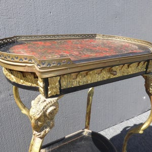 May include: A vintage, ornate side table with a black and gold finish. The table top is made of wood and has a red and gold inlay. The legs are carved in the shape of rams' heads.