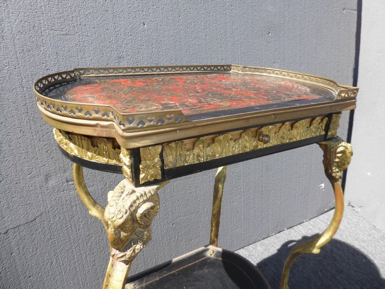 May include: A vintage, ornate side table with a black and gold finish. The table top is made of wood and has a red and gold inlay. The legs are carved in the shape of rams' heads.