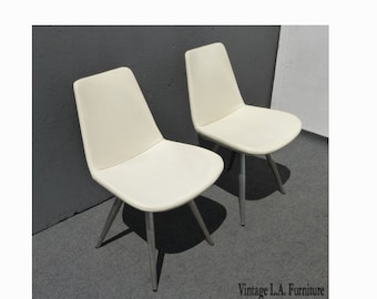 May include: Two white dining chairs with silver metal legs. The chairs have a simple, modern design.