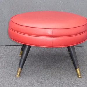 May include: A red round ottoman with black legs and gold accents. The ottoman has a tufted design and is upholstered in a smooth fabric.