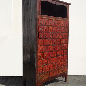 Op de afbeelding: Antiek Chinees rood en zwart houten kast met meerdere kleine laden. De kast heeft een klein open vak bovenaan en een paar grotere laden onderaan.