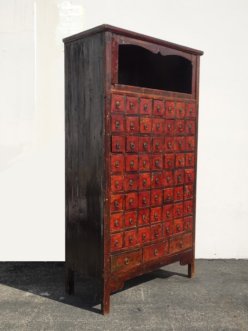 Op de afbeelding: Antiek Chinees rood en zwart houten kast met meerdere kleine laden. De kast heeft een klein open vak bovenaan en een paar grotere laden onderaan.