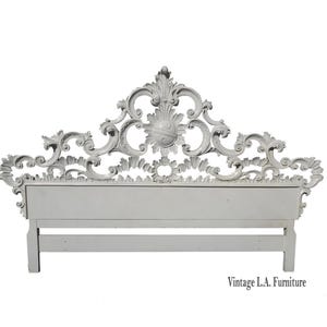 May include: White ornate headboard with a carved design. The headboard is made of wood and has a white finish. The headboard is attached to a wooden frame with two vertical supports.