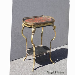 May include: A gold-tone metal side table with two tiers. The top tier has a red and black oval-shaped top with gold accents. The bottom tier is black with gold accents. The table has four legs with ornate details.