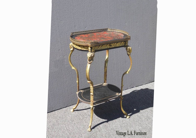 May include: A gold-tone metal side table with two tiers. The top tier has a red and black oval-shaped top with gold accents. The bottom tier is black with gold accents. The table has four legs with ornate details.