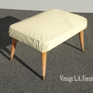 May include: A white upholstered ottoman with wooden legs and gold accents. The ottoman is sitting on a gray carpet.