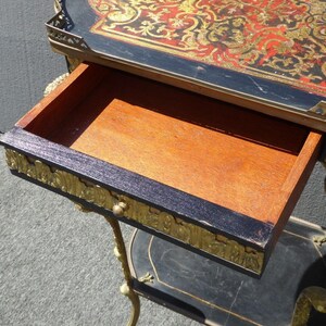 May include: A black and gold side table with a drawer and a shelf. The top of the table is decorated with a red and gold marquetry design.