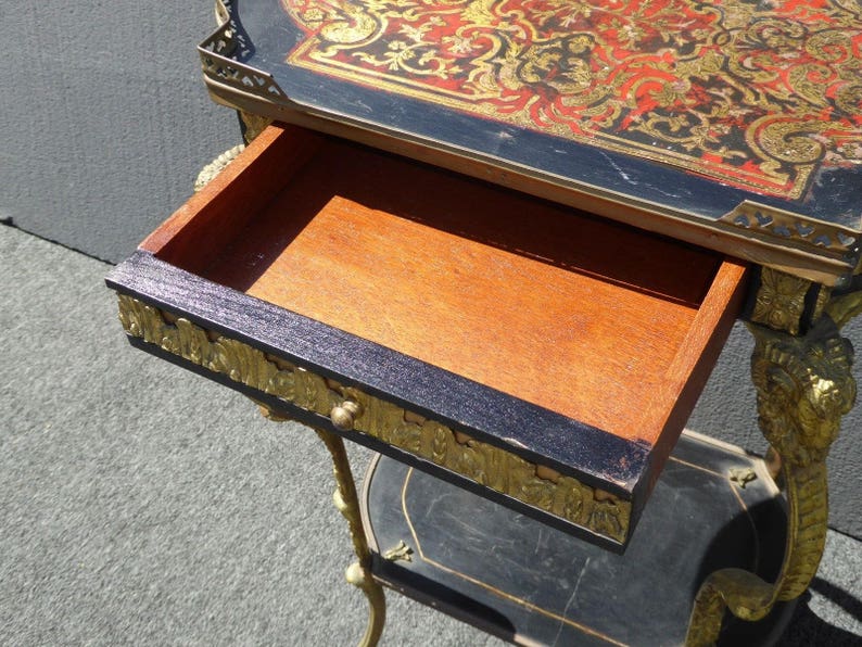 May include: A black and gold side table with a drawer and a shelf. The top of the table is decorated with a red and gold marquetry design.