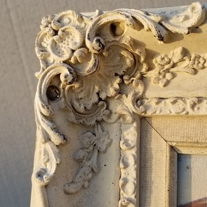 May include: Close-up of a white ornate picture frame with intricate details and a textured surface. The frame is made of plaster or wood and has a distressed finish.