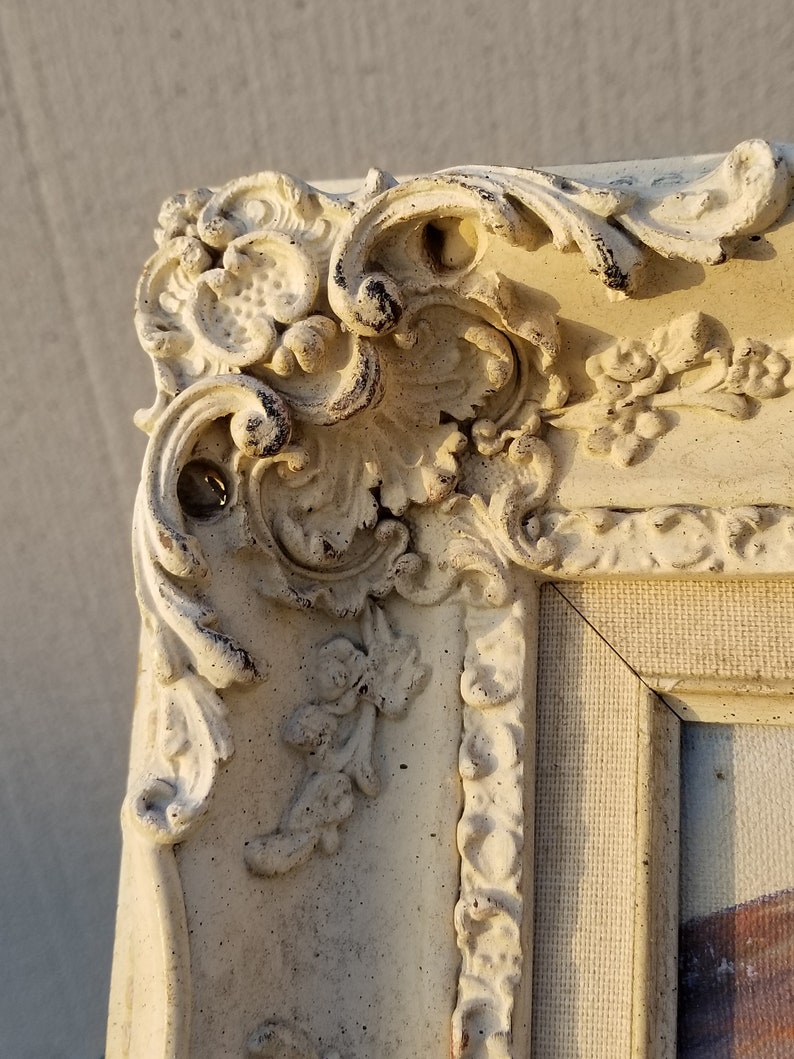 May include: Close-up of a white ornate picture frame with intricate details and a textured surface. The frame is made of plaster or wood and has a distressed finish.