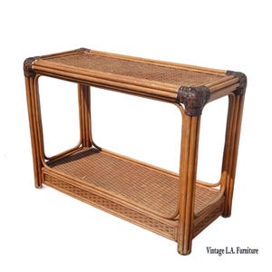 May include: A vintage rattan console table with two shelves. The table has a rectangular top and bottom shelf with a woven pattern. The corners are reinforced with darker brown accents. The table is made of light brown rattan.