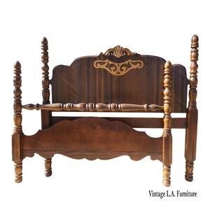 May include: A vintage wooden bed frame with a dark brown finish. The headboard features ornate carvings and a decorative gold-colored accent. The bed has four tall, turned posts and a footboard with a curved design. The text "Vintage L.A. Furniture" is at the bottom.
