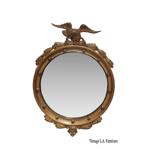 May include: A round, gold-colored mirror with an eagle perched at the top. The mirror has a decorative frame with a series of small, round accents. The bottom of the frame features a decorative floral design. The text "Vintage L.A. Furniture" is at the bottom.