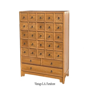 May include: A vintage wooden apothecary cabinet with multiple small drawers and larger drawers at the bottom. Each drawer has a dark ring pull. The cabinet is a light brown color and has a floral design on the drawer fronts. The text "Vintage L.A. Furniture" is at the bottom.