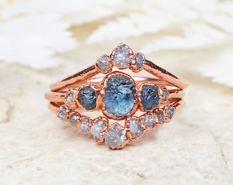 May include: A set of three rings with a rose gold finish. The rings feature light blue and clear gemstones. The central ring has an oval-shaped blue stone, while the other rings have smaller, clear stones. The rings are displayed on a textured white surface.