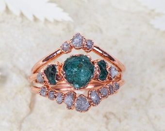 May include: A set of three rose gold rings with a green gemstone and small clear gemstones. The rings are arranged in a V-shape.