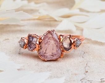 May include: A rose gold ring featuring a cluster of raw, uncut gemstones. The central stone is a large, light pink crystal, flanked by smaller, gray and purple stones. The ring is set against a neutral background with white floral accents.