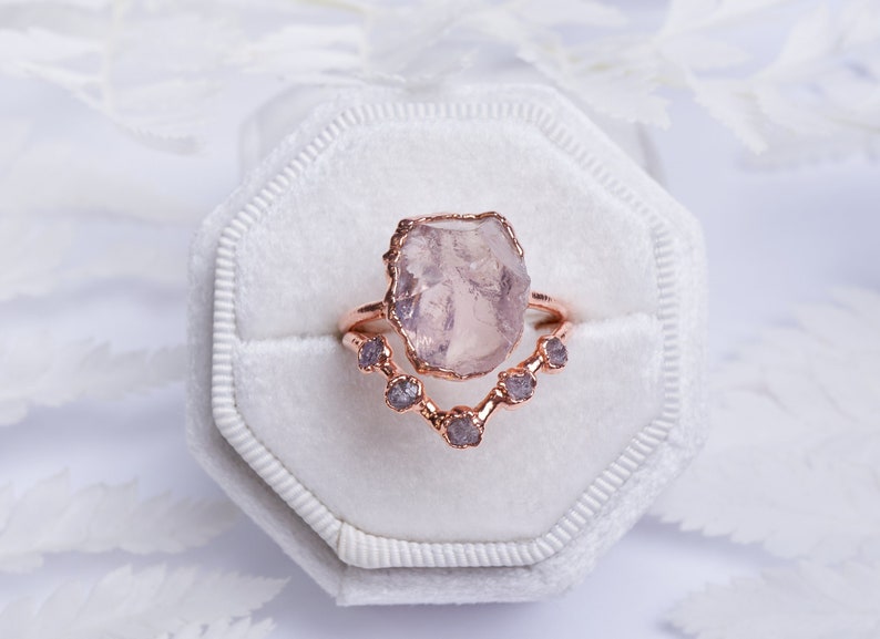 May include: A rose gold ring set with a large, light pink, raw, irregular shaped gemstone. The ring is set with a smaller row of five, clear, raw, irregular shaped gemstones.