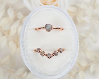 May include: Two rose gold rings featuring raw, uncut grey diamonds. The top ring has a single central stone, while the bottom ring is V-shaped and set with five small, irregular grey diamonds along the band, displayed in a white velvet ring box.