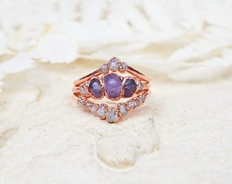 May include: A rose gold ring set featuring a central band with three raw purple stones and two outer bands adorned with small, rough-cut white diamonds. The metal has a warm, metallic finish, and the stones are set in a prong-style arrangement.