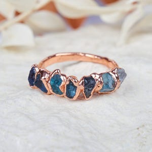 May include: A rose gold ring featuring a series of raw, uncut blue gemstones set along the top half of the band. The stones vary in shades of deep navy, teal, and light blue, each held in place by a textured, metallic rose gold setting.
