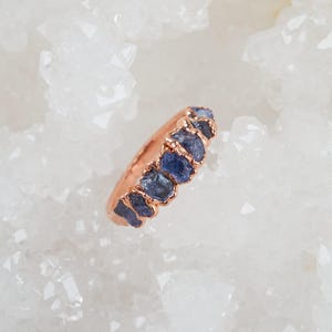 May include: A rose gold ring featuring a row of five small, rough-cut blue sapphire gemstones set in individual prongs. The band has a smooth, polished finish and the stones exhibit a natural, irregular texture with deep blue hues.