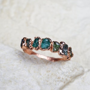 Puede incluir: Un anillo de oro rosa con una hilera de siete piedras preciosas en bruto, sin tallar, de color azul verdoso, engastadas en una banda de metal texturizado. Las piedras varían en tamaño y forma, mostrando tonos azul verdosos profundos.
