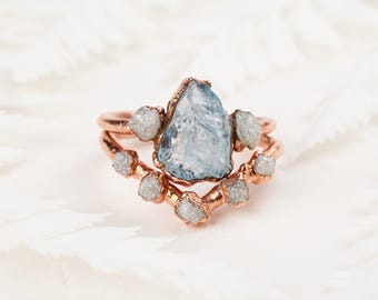 May include: A rose gold ring set featuring a central pear-shaped raw blue gemstone flanked by two small white raw stones, paired with a curved band adorned with seven small white raw stones along the top edge of the metal band.