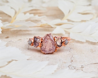 Puede incluir: Un anillo de oro rosa con una piedra preciosa rosa en bruto en forma de pera en el centro, flanqueada por dos piedras en bruto moradas más pequeñas y dos diminutas piedras grises a cada lado, sobre una banda de metal con bordes orgánicos.