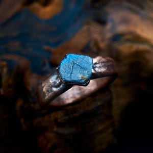 May include: A ring featuring a raw, hexagonal blue gemstone set into a textured, dark metallic band. The stone displays a vibrant blue hue with visible surface facets, while the band has a rugged, uneven finish with dark brown and copper tones.
