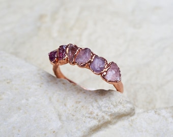 Può includere: Un anello color rame caratterizzato da una fila di sei pietre preziose grezze e non tagliate in varie tonalità di rosa e viola. Le pietre sono incastonate in una fascia metallica con una finitura organica e testurizzata.