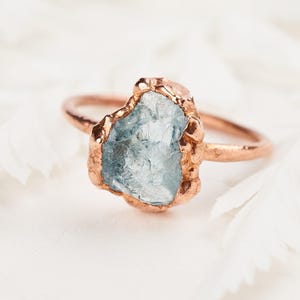 May include: A ring featuring a raw, light blue, translucent gemstone set in a textured, metallic copper-colored band. The stone has an irregular, jagged shape and is held in place by a thick, uneven metal setting that matches the band's finish.