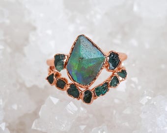 Puede incluir: Un anillo de oro rosa con una piedra central de ópalo grande, irregular e iridiscente con destellos verdes y azules, flanqueada por piedras más pequeñas, en bruto y de color verde azulado oscuro, engastadas en una banda curva en forma de V.