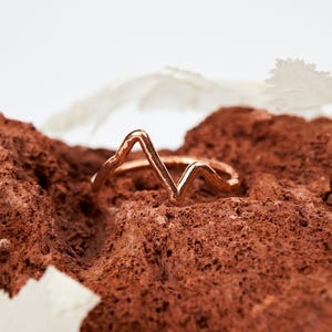 May include: A rose gold-toned ring featuring a continuous, angular mountain range silhouette. The metal band has a slightly hammered, textured finish, creating a distinct peak and valley pattern that wraps around the finger in a minimalist design.