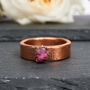 Puede incluir: Un anillo de cobre con una piedra preciosa de rubí en bruto engastada en el centro. El anillo tiene una textura martillada y un aspecto rústico.