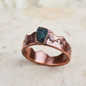 May include: A copper ring featuring a textured, uneven band with a mountain-like silhouette and a single, raw, dark green gemstone set prominently at the top. The metal surface has a polished, reddish-brown metallic finish with visible indentations.