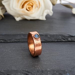 Puede incluir: Un anillo ancho de cobre con un acabado texturizado y martillado que presenta una pequeña piedra preciosa azul de forma triangular engastada en la banda. El metal tiene un brillo cálido de color marrón rojizo y una superficie irregular.
