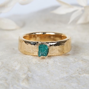 Puede incluir: Un anillo de tono dorado con una banda martillada y texturizada que presenta una única piedra pequeña, cruda y de color verde azulado engastada en el centro. La superficie metálica tiene un acabado orgánico irregular y la piedra es rugosa.