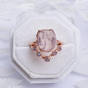 May include: A rose gold ring featuring a large, raw, pale pink crystal centrepiece. The band is shaped in a V-curve and is accented with five small, round, light purple stones set along the lower edge, creating a layered, textured appearance.